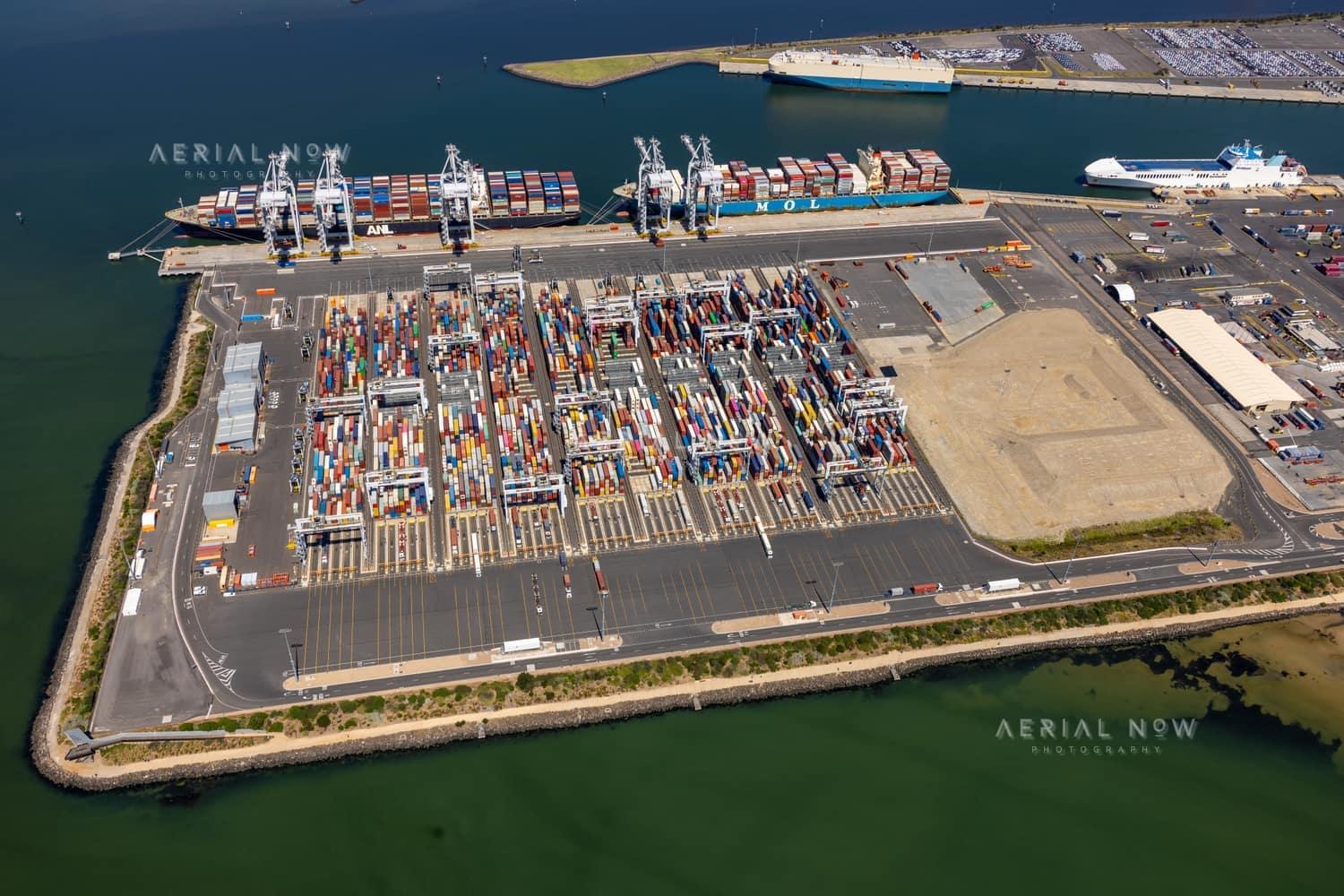 Aerial view of Webb Dock East at the Port of Melbourne in Port Melbourne, showing container stacks, ship-to-shore cranes, berthed container vessels, port roads and surrounding harbour water.