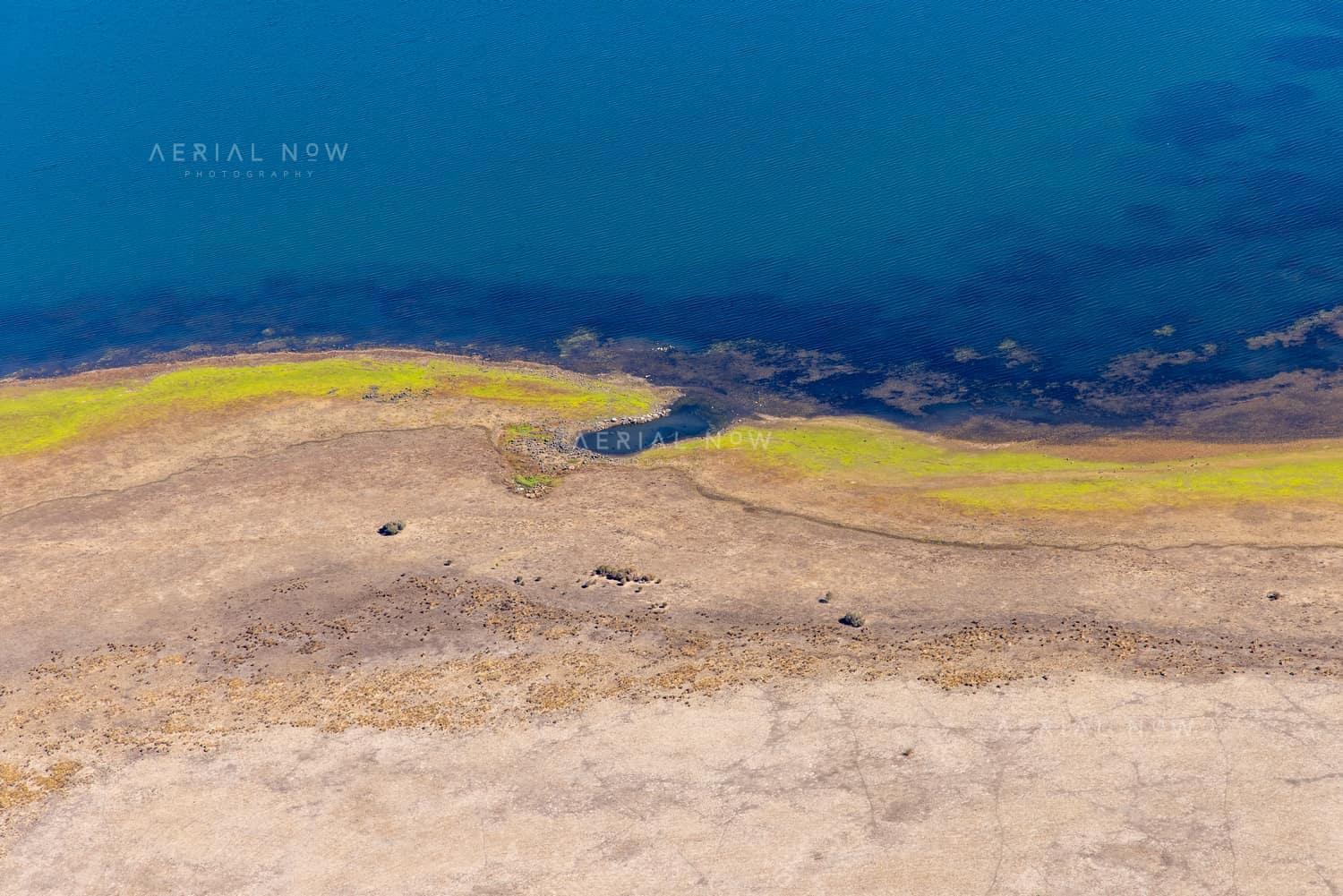 Aerial view of Lake Modewarre shoreline at Modewarre VIC, showing blue water, exposed lakebed and yellow-green vegetation along the shallow edge.