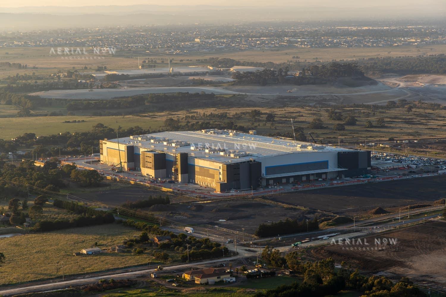 Aerial view of a large industrial warehouse complex surrounded by roads, car parks, open land and developing estates in Mickleham, Victoria.