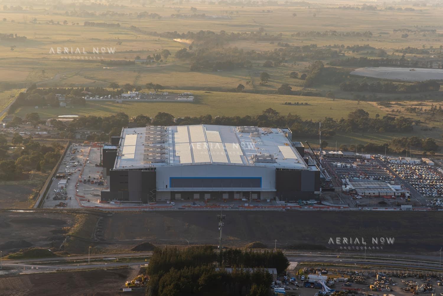 Aerial view of the Amazon Robotics Fulfilment Centre under construction in Craigieburn, with a large warehouse building, loading areas, staff parking and surrounding rural land.