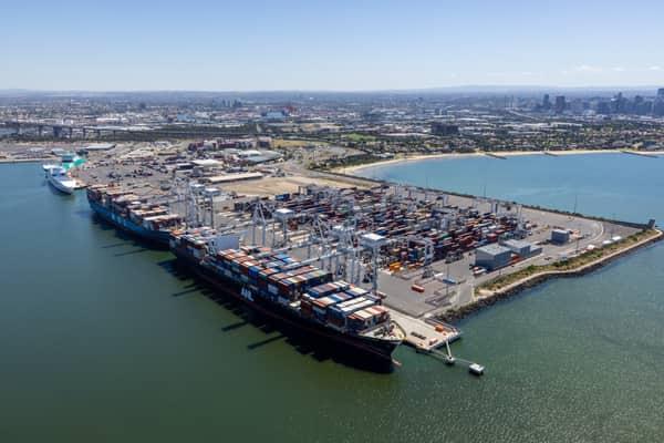 Aerial view of container ships berthed at Webb Dock East in Port Melbourne, with ship-to-shore cranes, container stacks, Port Phillip Bay and the Melbourne CBD skyline in the background.
