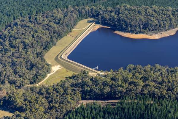 Aerial view of Bostock Reservoir at Spargo Creek, showing the earth embankment, access road, outlet structure, dark water and surrounding forest.