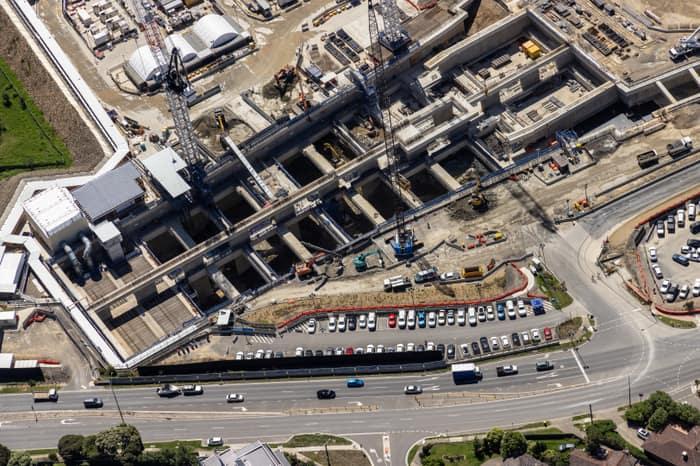 Aerial view of North East Link construction works in Bulleen, showing concrete tunnel and road structures, cranes, work zones, parked vehicles and traffic on the adjoining arterial road.