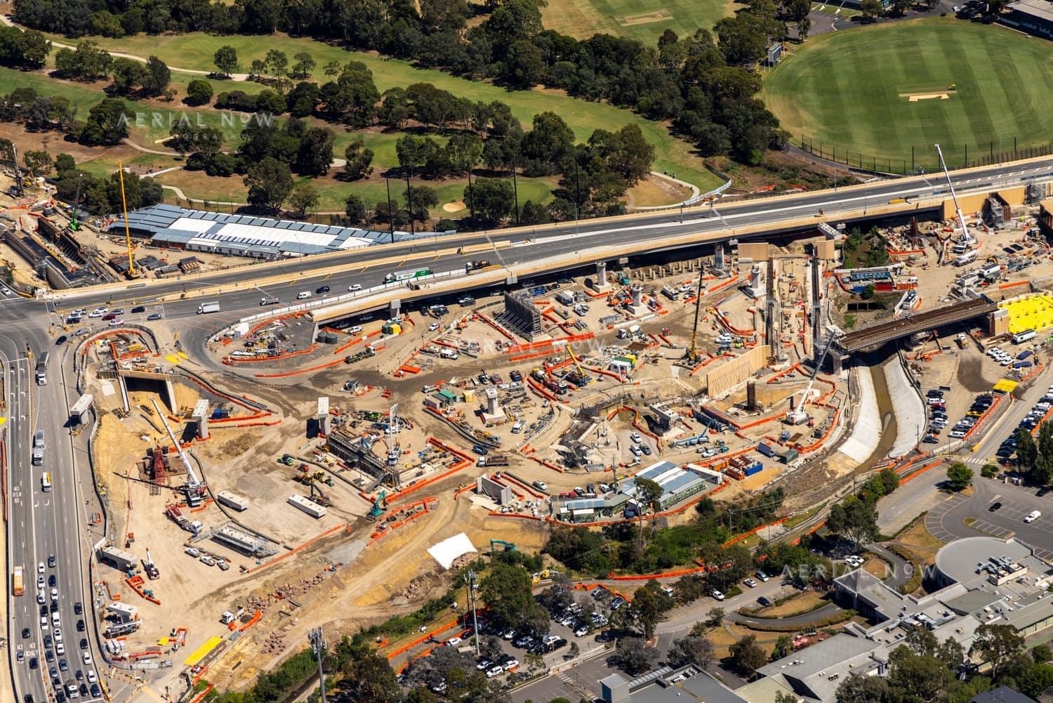 Aerial view of the North East Link construction site beside Manningham Road in Bulleen, with cranes, bridge works, traffic, parkland and a nearby sports oval visible.