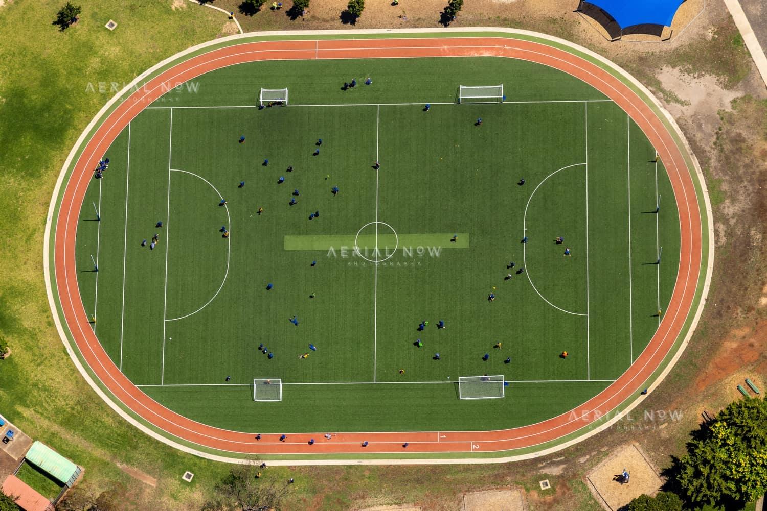 Aerial view of a soccer field with goalposts, player groups and a surrounding red athletics track near Astor Place in Melton West, Victoria.
